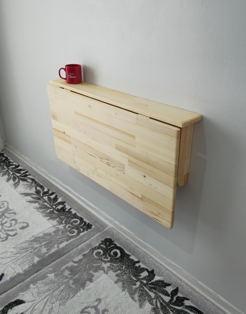 May include: A light-coloured, wall-mounted, folding table made of wood. A red mug with white text sits on the table's surface. The table is mounted on a light grey wall, above a patterned rug.