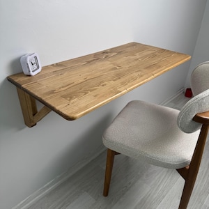 May include: A wall-mounted wooden desk with a natural finish. The desk is supported by a triangular bracket and is paired with a light-coloured upholstered chair. A small white alarm clock sits on the desk.