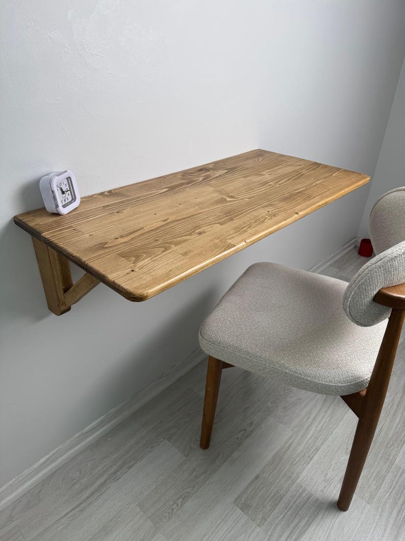 May include: A wall-mounted wooden desk with a natural finish. The desk is supported by a triangular bracket and is paired with a light-coloured upholstered chair. A small white alarm clock sits on the desk.