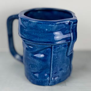 May include: A ceramic mug with a denim design. The mug is a deep blue color, with details that mimic the look of a pair of jeans, including seams and pockets. The mug has a curved handle and a glossy finish.