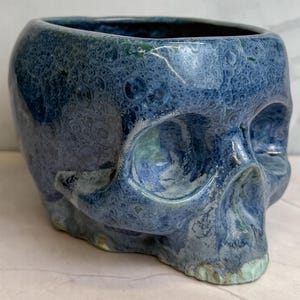 May include: A ceramic skull-shaped planter featuring a glossy, crystalline glaze in shades of deep blue, light blue, and hints of green. The surface has a textured, mottled appearance with visible circular patterns throughout the blue finish.