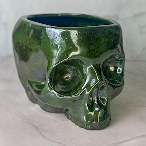 May include: A skull-shaped ceramic vessel with a glossy, deep forest green glaze. The object features a hollow top opening, defined eye sockets, a nasal cavity, and a detailed jawline, all finished with a reflective, metallic-like surface texture.
