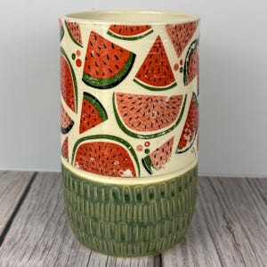 May include: A ceramic vase with a watermelon design. The top portion features red watermelon slices with green rinds on a cream background. The bottom half is a textured green. The vase is cylindrical.