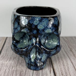 May include: A ceramic skull-shaped planter with a square top. The planter is glazed in shades of blue and green, with a speckled pattern. The interior is black. The skull has detailed features and sits on a wooden surface.