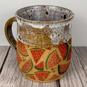 May include: A handmade ceramic mug with a watermelon design. The mug features a brown handle and a speckled, off-white rim. The lower half is decorated with red watermelon slices with green rinds on a tan background. A unique, functional piece.