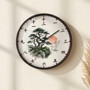 May include: A round wall clock with a black frame and a white face featuring a painted illustration of a green pine tree on a rock, bamboo stalks, and a red sun. The hour markers are represented by black Japanese kanji numerals from one to twelve.