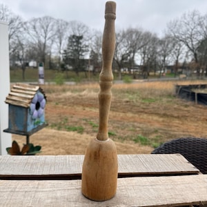 May include: A vintage wooden butter churn with a long, turned handle and a rounded base. The churn is made of light-colored wood and sits on a rustic wooden surface. The background features a birdhouse and a natural outdoor setting.