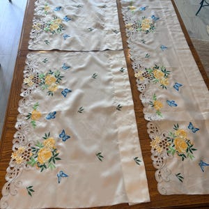 May include: Three cream-colored fabric panels featuring intricate cutwork edges and embroidered floral motifs. The design includes clusters of yellow roses with green leaves and small blue butterflies scattered across the smooth textile surface.