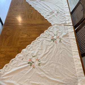May include: Two off-white curtain panels featuring a scalloped edge with embroidered scrollwork and floral motifs in shades of coral, white, and green. The fabric is semi-sheer with a straight top hem and a diagonal bottom edge with embroidery.