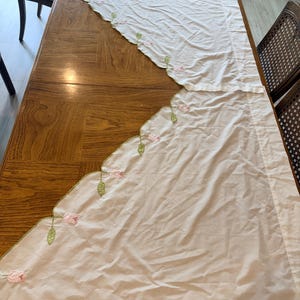 May include: A pair of off-white window curtains featuring a scalloped edge with embroidered pink rosebuds and green leaves. The fabric is lightweight and semi-sheer, with a straight top hem and a decorative floral pattern along the curved bottom.