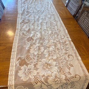 May include: A long, rectangular white lace table runner featuring an intricate floral pattern with detailed petals and leaves. The fabric has a sheer, open-weave mesh texture with a decorative border along the edges, showcasing a delicate design.