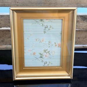 May include: A rectangular picture frame with a metallic gold finish and a beveled border. The interior display features a light blue background with horizontal lines and a delicate pattern of small pink flowers and green leaves.