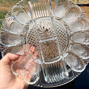 May include: Clear glass serving platter with multiple compartments. The platter has a scalloped edge and a central section with a diamond pattern. The compartments are shaped like shells. A hand is holding the platter.