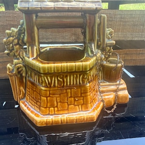 May include: A glazed ceramic wishing well planter in shades of brown and green. The structure features a stone-textured base with the word WISHING embossed on the front, a roofed top, and a small attached bucket suspended by a thin metal chain.