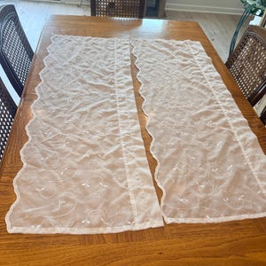 May include: Two rectangular sheer white curtain panels featuring a delicate embroidered vine pattern with small leaves. Each panel has a scalloped edge along the side and a straight hem at the top, crafted from a lightweight, semi-transparent fabric.