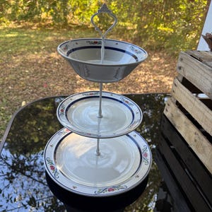 May include: A three-tiered serving stand with white ceramic plates and a bowl. The plates and bowl have a blue border with floral accents. The stand is made of white metal and has a decorative handle.