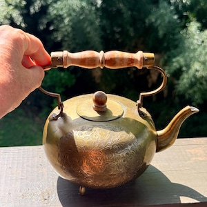 May include: A vintage brass teapot with a wooden handle and lid knob. The teapot has an etched floral design and sits on three small feet. The handle is held by a hand. The teapot is on a wooden surface.