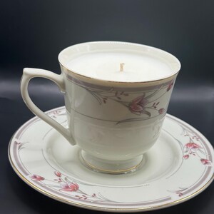 May include: A white teacup candle with a floral design, resting on a matching saucer. The teacup and saucer have a pink and gray floral pattern with gold trim. The candle wax is white, and a single wick is visible.