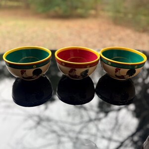 May include: Three ceramic bowls with a rooster design. Each bowl has a different interior color: green, red, and green. The rims are yellow, and the exterior is cream-colored. The bowls are arranged in a row, reflecting on a dark surface.
