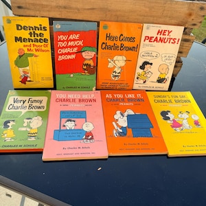 May include: A collection of vintage comic books featuring characters from the Peanuts and Dennis the Menace series. The books have colorful covers with titles like "Here Comes Charlie Brown!" and "Dennis the Menace and Poor Ol' Mr. Wilson."
