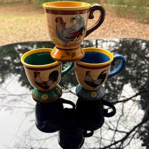 May include: Three decorative pedestal mugs with rooster designs. The mugs have a tan base color with a rooster illustration in the center. Each mug has a different colored interior and handle: green, blue, and brown. The mugs are stacked on top of each other.