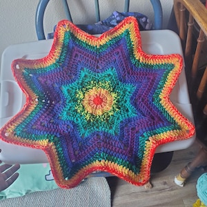 May include: A handmade, rainbow-colored crochet star. The star features a vibrant spectrum of colors, including red, orange, yellow, green, blue, and purple. The star's design is intricate, with a central circular pattern and pointed edges.