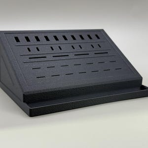 May include: A dark grey display stand with a slanted top featuring various slots and cutouts. The stand has a textured finish and a small tray at the bottom. It appears designed for displaying small objects.