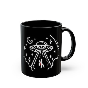 May include: Black ceramic mug with a white illustration of a UFO beaming up a stick figure. The design includes stars, clouds, and a crescent moon. The mug has a curved handle.