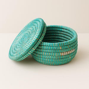 May include: A turquoise woven basket with a lid. The basket and lid are made of tightly woven, light-coloured material with turquoise accents. The lid is partially open, revealing the interior of the basket. The basket is round.