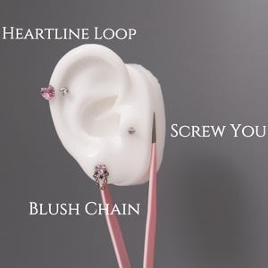 May include: A white ear model with three pieces of jewellery: a heart-shaped stud, a small stud, and a chain earring. The text "HEARTLINE LOOP", "SCREW YOU", and "BLUSH CHAIN" are visible.