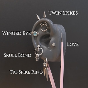 Op de afbeelding: Een display van verschillende oorpiercings op een grijs oor model. De piercings omvatten een gevleugelde oogstud, een skull bond-hoepel, een tri-spike ring en twin spikes. Het woord "LOVE" is ook zichtbaar.