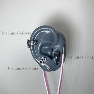 May include: A gray ear model with silver-colored jewelry, including a cuff, scissors, and a zipper-shaped stud. The text "The Tailor's Zipper", "The Tailor's Shears", and "The Tailor's Pull" are visible. Pink tweezers are positioned near the ear.