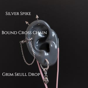 May include: A gray ear model displays multiple silver piercings. The piercings include silver spikes, a bound cross chain, and a grim skull drop. The text "SILVER SPIKE", "BOUND CROSS CHAIN", and "GRIM SKULL DROP" are visible.
