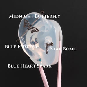 Könnte beinhalten: Ein Silikon-Ohrmodell mit vier Schmuckstücken: ein dunkler industrieller Metallstab mit Schmetterlingsanhänger namens Midnight Butterfly, ein blauer Schmetterlingsstecker namens Blue Flutter, ein Knochen- und Sternanhänger namens Star Bone und ein blauer Herzstecker namens Blue Heart Spark.