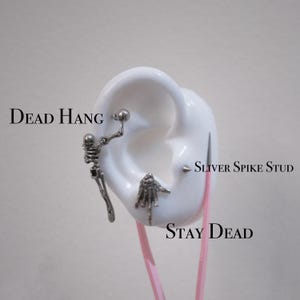 Puede incluir: Exhibición de tres pendientes plateados. Uno presenta un diseño de esqueleto, otro una mano y el tercero es un pendiente de punta. Las palabras "Dead Hang", "Sliver Spike Stud" y "Stay Dead" están impresas en la imagen.