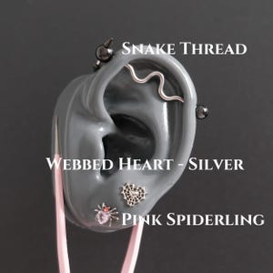May include: A gray ear model displaying three piercings: a silver snake thread industrial barbell, a silver webbed heart stud, and a pink spiderling stud with a heart-shaped gemstone. The text labels read SNAKE THREAD, WEBBED HEART - SILVER, and PINK SPIDERLING.