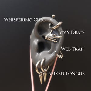 May include: A display of silver-colored ear piercings on a gray ear model. The piercings include a spiral, a skeleton hand, a web-like design, and hoop earrings with cone-shaped spikes. Text on the image reads "WHISPERING CURL", "STAY DEAD", "WEB TRAP", and "SPIKED TONGUE".