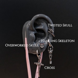 Può includere: Esposizione di orecchini argentati, tra cui un teschio contorto, uno scheletro appeso, una croce e un teschio sovraccaricato. Gli orecchini sono esposti su un modello di orecchio grigio con il testo "TWISTED SKULL", "HANGING SKELETON", "OVERWORKED SKULL" e "CROSS".