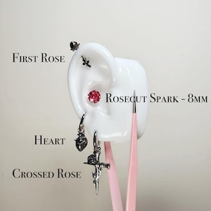 Puede incluir: Una variedad de pendientes y tachuelas plateados exhibidos en un modelo de oreja blanco. Los pendientes incluyen un dije en forma de corazón, una cruz con una rosa y un pequeño arete de rosa. También se presenta un arete rojo con corte de rosa, etiquetado como "Rosecut Spark - 8mm".