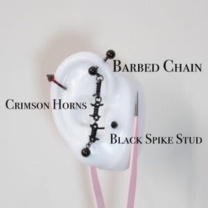 May include: An ear model displays several pieces of body jewelry. A crimson-colored horn-shaped earring, a black barbell, a black barbed chain earring, and a black spike stud are visible. The text "Barbed Chain", "Crimson Horns", and "Black Spike Stud" are also present.