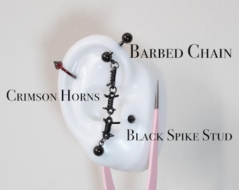 Könnte beinhalten: Ein Ohrmodell zeigt verschiedene Körperschmuckstücke. Ein karmesinroter, hornförmiger Ohrring, eine schwarze Langhantel, ein schwarzer Ohrring mit Stacheldrahtkette und ein schwarzer Spikestift sind sichtbar. Der Text "Barbed Chain", "Crimson Horns" und "Black Spike Stud" ist ebenfalls vorhanden.