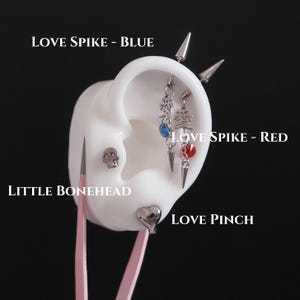 May include: A white ear model displaying multiple silver-toned piercings: two industrial-style bars with spikes and dangling charms featuring Japanese characters and blue or red crystals, a skull stud labeled Little Bonehead, and a heart stud labeled Love Pinch.