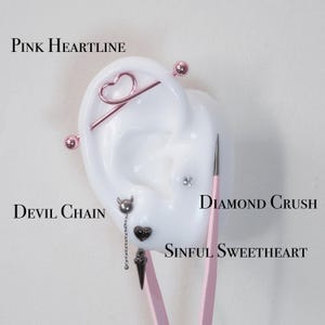 May include: A selection of ear piercings presented on a white ear model. The piercings include a pink heart-shaped barbell, a silver devil chain, a diamond stud, and a black heart stud. The text "Pink Heartline", "Devil Chain", "Diamond Crush", and "Sinful Sweetheart" are also visible.