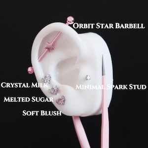Puede incluir: Una exhibición de piercings para la oreja, incluyendo un Orbit Star Barbell, Crystal Milk, Melted Sugar y Soft Blush. También se muestra el Minimal Spark Stud. La joyería es rosa y plateada, sobre un fondo negro.