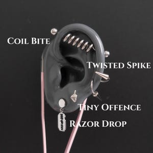 Könnte beinhalten: Ein graues Ohrmodell mit vier silberfarbenen Piercings, beschriftet mit: Coil Bite, ein spiralförmiger Stab; Twisted Spike, ein gebogener Dorn; Tiny Offence, ein Stecker in Handform; und Razor Drop, ein Rasierklingen-Anhänger. Metallschmuck.