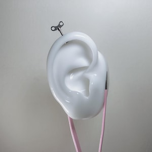 May include: A white ear sculpture with a wind-up key inserted into the top. Pink tweezers are positioned below the ear. The image is set against a neutral gray background.