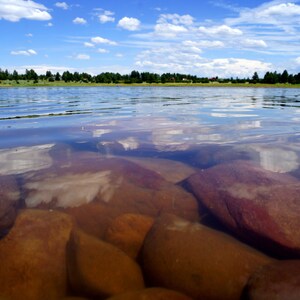 May include: A clear blue lake with a bright blue sky and white clouds. The water is shallow and you can see large brown rocks at the bottom of the lake.