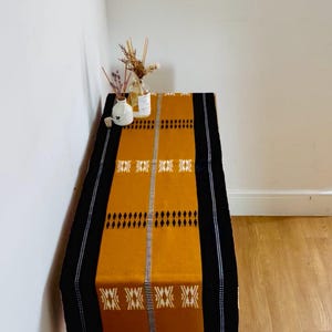 May include: A mustard yellow and black patterned table runner. The runner has a central mustard section with black and white geometric designs, framed by black borders. Decorative items are placed on the table, adding to the aesthetic.