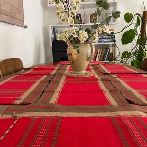 Op de afbeelding: Een rood en bruin geruite tafelkleed bedekt een houten tafel. Een beige keramische kan met bloemmotieven bevat witte bloemen. De tafel is gedekt met bijpassende rode en bruine placemats. Een wereldkaart hangt op de achtergrond aan de muur.
