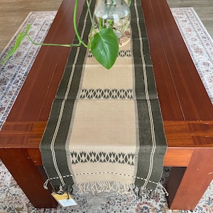 May include: A rectangular table runner with a central beige section and dark green borders. The runner features a geometric pattern and fringe at the ends. A glass vase with a green plant sits on the table, enhancing the home decor.